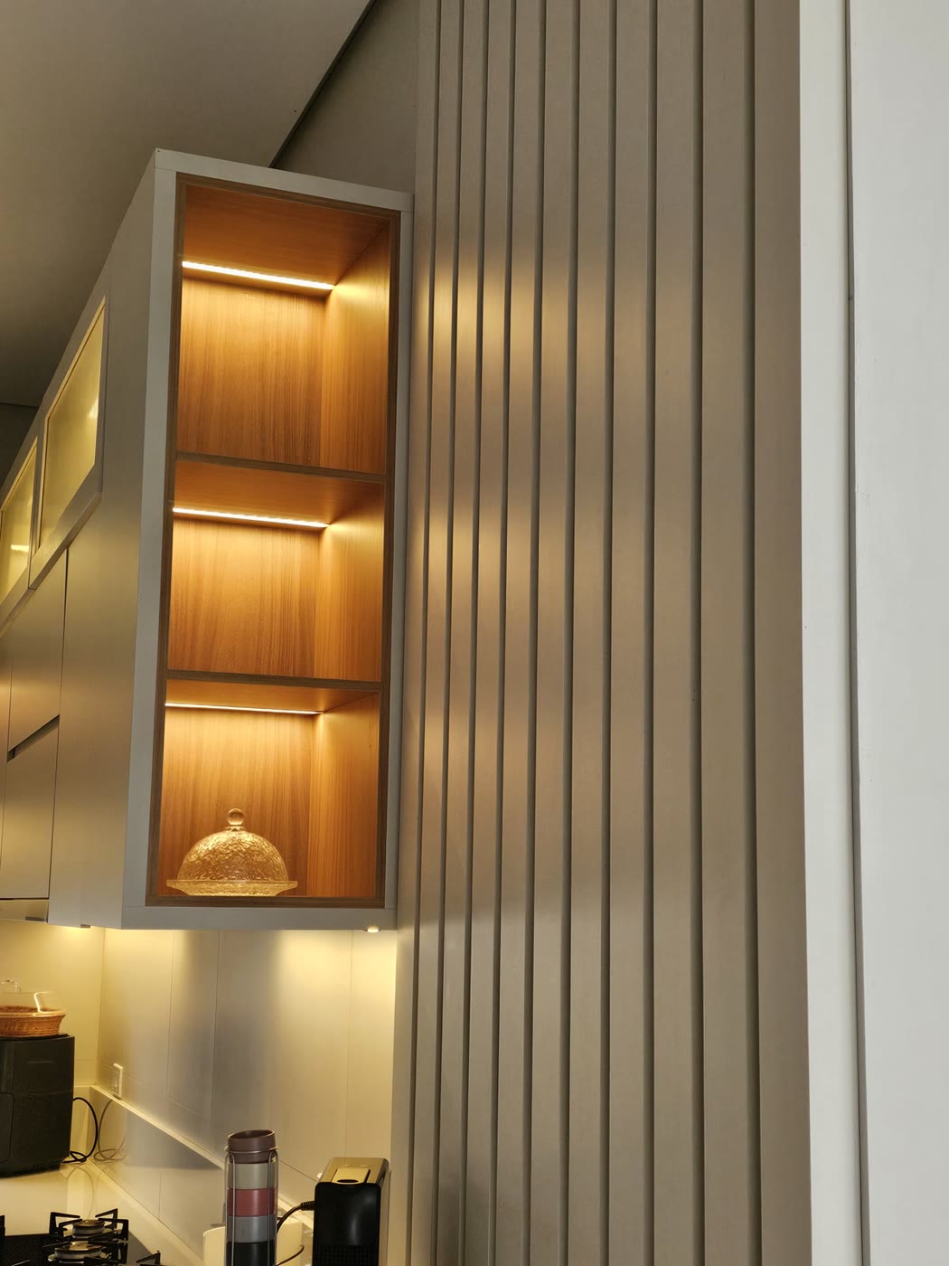 Nicho vertical de madeira com três prateleiras, cada uma com fita de LED acesa, ao lado de um painel ripado bege.