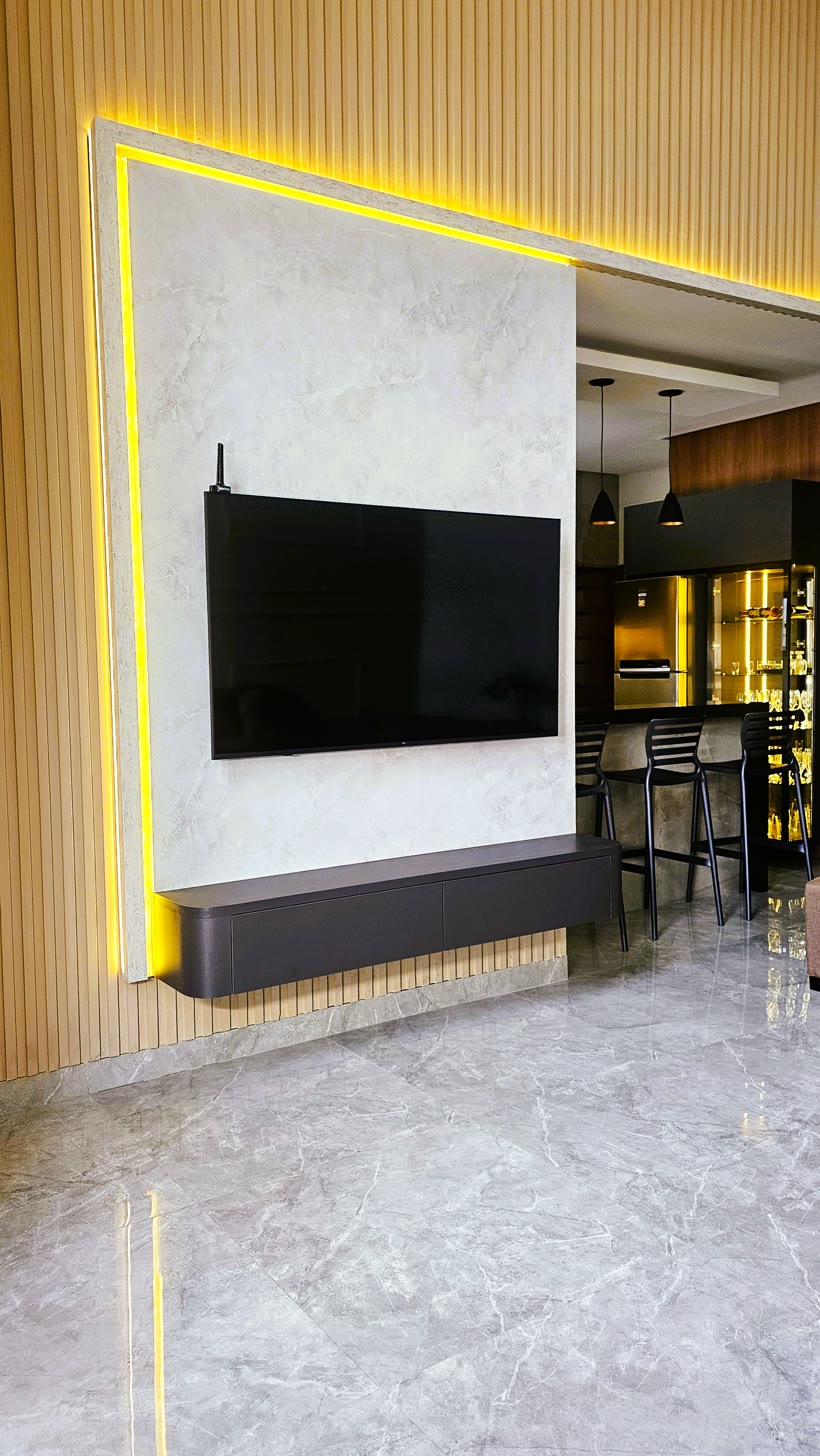 Painel de TV revestido em pedra clara com moldura de LED acesa em volta, sobre parede ripada de madeira.