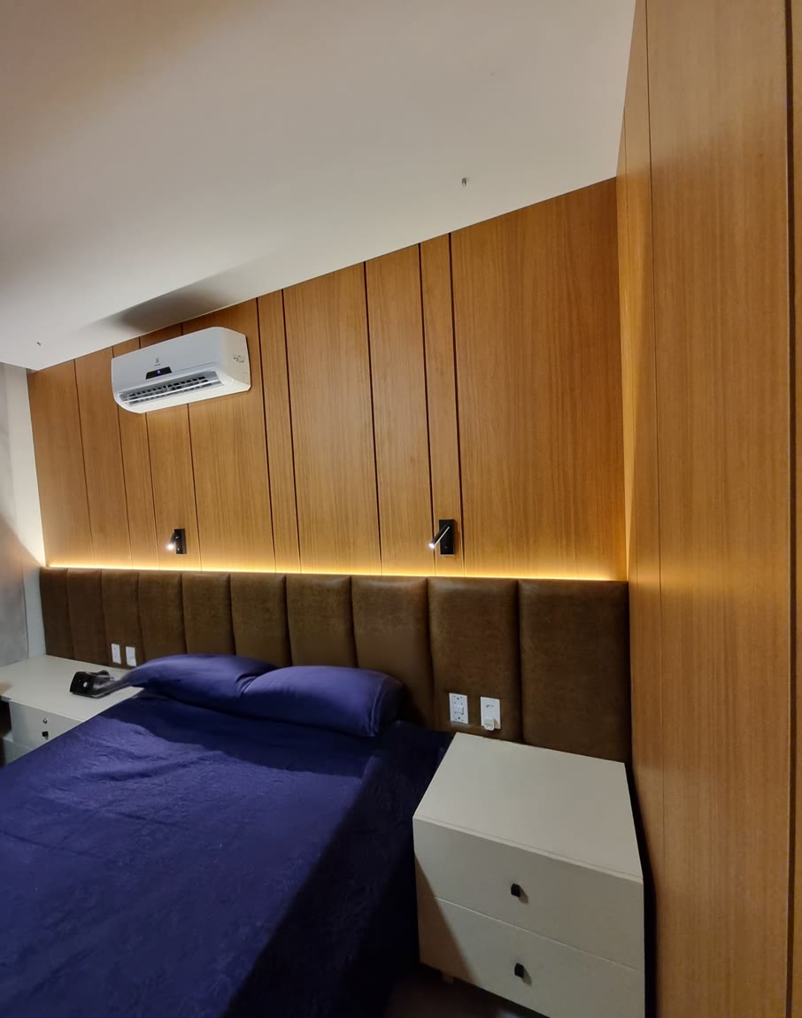 Quarto com parede inteira de armários em madeira, cabeceira estofada marrom e fita de LED acesa por cima.