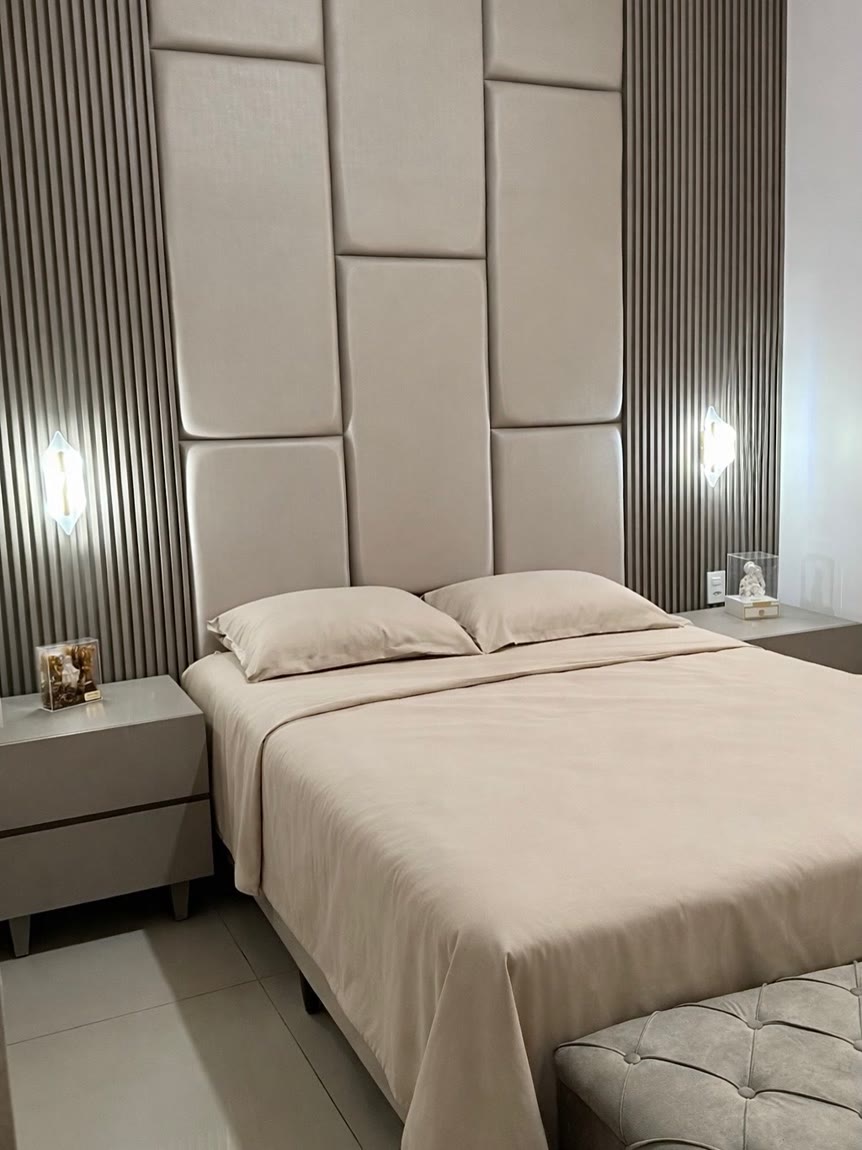 Quarto com cabeceira estofada em painéis e ripado escuro nas laterais, com arandelas acesas.