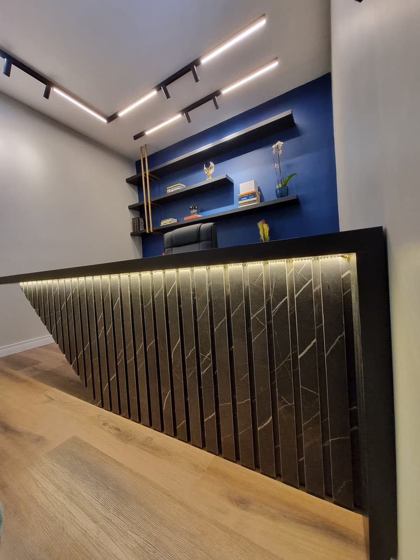 Recepção de escritório com balcão ripado em pedra e fita de LED por dentro, parede azul com prateleiras pretas.